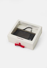 Fekete cipzáras tasak geometrikus mintával, arany hardware-rel, fehér dobozban átlátszó fedéllel és piros szalaggal bemutatva. Calvin Klein logó látható.