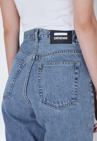 Modré džíny s vysokým pasem s zadní štítkem. Vyznačují se dvěma zadními kapsami, viditelným prošíváním a lehce vybledlou texturou.