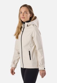 Ženska v svetlo bež jakni s kapuco, z črnimi zadrgami in detajli na žepih, stoji pred enobarvnim belim ozadjem.