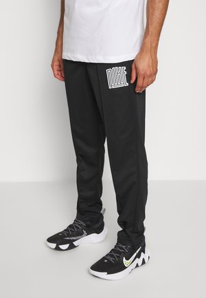 Mann trägt schwarze Nike-Sweathosen und schwarz-weiße Nike-Sneaker und steht auf einem hellen Boden vor einer weißen Wand.