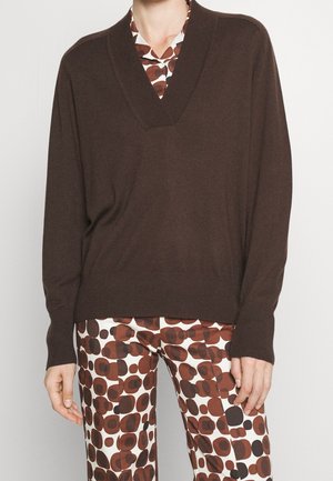 Personne portant un pull marron à col en V par-dessus une chemise à motifs et un pantalon assorti avec des formes circulaires marron et noires sur fond blanc.