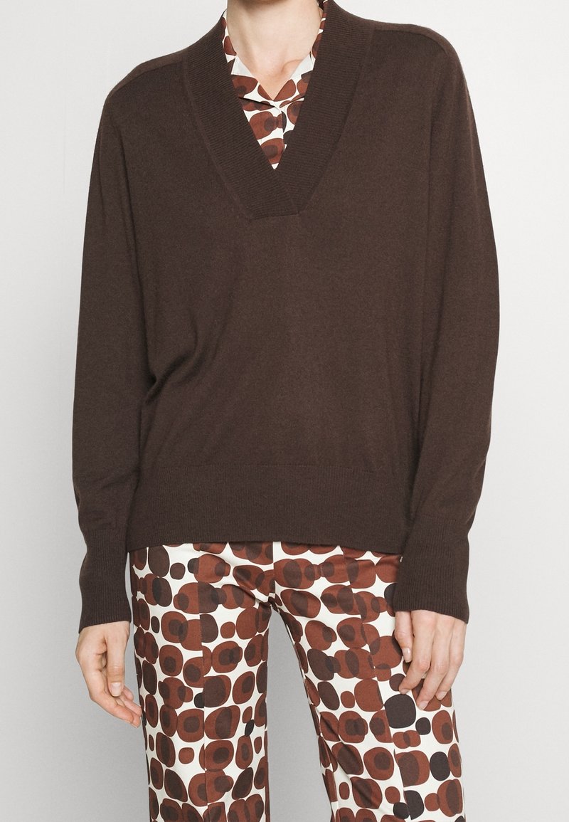 Personne portant un pull marron à col en V par-dessus une chemise à motifs et un pantalon assorti avec des formes circulaires marron et noires sur fond blanc.