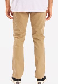 Chinos de algodón beige con un diseño de pierna recta, que presentan una textura suave y detalles de costura sutiles, combinados con zapatillas negras.
