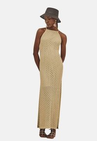 Vestido largo y ajustado con un patrón de punto de diamante en color tostado. Presenta un escote halter y aberturas laterales. Combinado con un sombrero de cubo negro de tejido.
