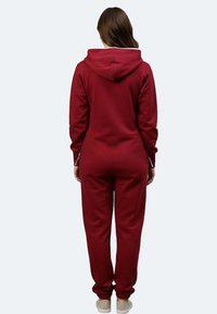 Bordeauxfarbener Kapuzenoverall mit langen Ärmeln, Kapuze mit Kordelzug und enganliegenden Bündchen. Verfügt über eine Reißverschlusstasche auf der Rückseite. Weicher, texturierter Stoff.