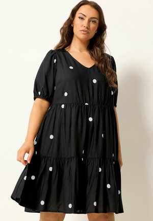 Kobieta ubrana w czarną sukienkę z falbanami w białe groszki, z krótkimi bufiastymi rękawami i dekoltem w serek, stojąca na jasnym tle.