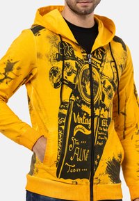 Sweat à capuche zippé jaune avec design graphique noir représentant une moto, texture vieillie et accents noirs contrastants.