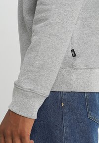 Ingrandimento di una persona che indossa una felpa grigia dei Vans e jeans blu, mostrando il dettaglio del maniche della felpa e del tag.