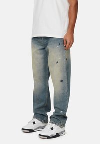 Blauwe denim jeans met een vervaagde afwerking, voorzien van verfspatten in lichtblauw en zwart. Rechte pijpen, gedragen met witte sneakers.