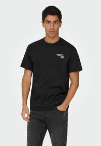 Camiseta de algodón negra con cuello redondo y mangas cortas, que presenta un estampado gráfico blanco en el lado izquierdo del pecho. Corte holgado, textura suave.