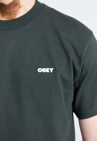 Dark green cotton t-shirt with crew neck. Features white "OBEY" logo printed on the left chest area. Smooth texture and casual fit.