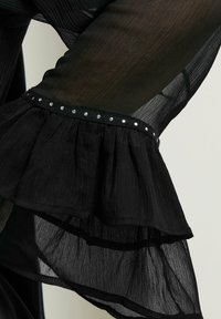 Manica in tessuto nero trasparente con un design arricciato. Presenta una banda orizzontale accentuata da borchie argentate lungo il bordo. Texture leggera.