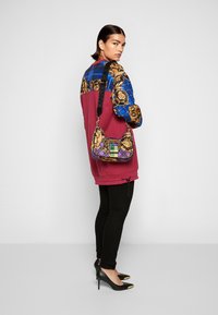 Borsa a tracolla con motivi colorati, caratterizzata da dettagli dorati e una base viola. Modella che indossa un top color bordeaux e leggings neri, con tacchi a punta neri.