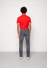 Red polo shirt with white accents, paired with dark gray slim-fit jeans. Back view shows a smooth texture and fitted design. White sneakers.