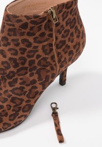 Stivale alla caviglia in suede marrone con stampa leopardata e tacco a stiletto nero. Include una cinghia removibile con clip metallica.