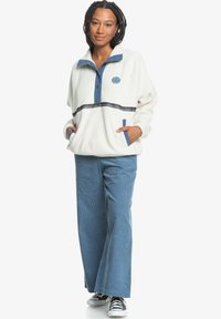 Weißer Fleece-Pullover mit blauen Akzenten, Druckknöpfen und einem Logo auf der Vorderseite. Kombiniert mit blauen Cordhosen und schwarz-weißen Turnschuhen.