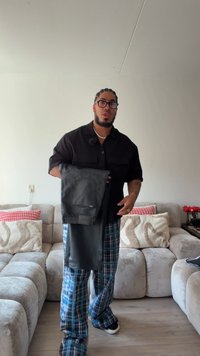 Zwarte leren broek met een getailleerde fit, voorzien van een achterzak en een gladde textuur. Gepresenteerd door een persoon in een zwart shirt en geruite broek.