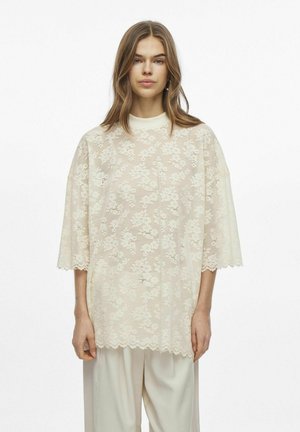 Blusa de encaje crema con patrones florales, de corte holgado, mangas hasta el codo, dobladillo ondulado, llevada sobre pantalones beige holgados.