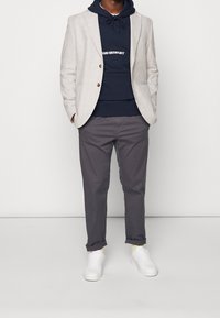 Ljusgrå rutad blazer över en marinblå hoodie med vit text, tillsammans med mörkgrå byxor och vita sneakers.