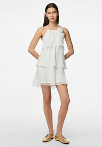 Robe blanche sans manches avec trois volants superposés, un col halter et un accent floral en tissu à l'épaule ; texture lisse.
