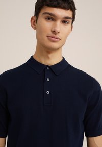 Tums zils īsām piedurknēm polo krekls ar apkakli, trijām pogām un mīkstu, gludu tekstūru.