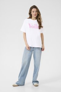 T-shirt blanc en coton avec le mot "Voilà" en lettres roses, associé à un jean bleu clair ample et des chaussures slip-on beiges. L'arrière-plan est uni.