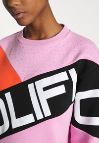 Sudadera colorida con segmentos en rosa, naranja y negro, acentos de strass y letras blancas en negrita en el pecho.