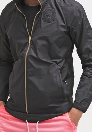 Schwarze Jacke mit Reißverschluss und rundem Logo auf der Brust, getragen über einem schwarzen Shirt, kombiniert mit pinken Hose mit Kordelzug. Hände in den Taschen.