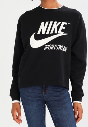 Persoon die een zwarte Nike Sportswear trui en een blauwe spijkerbroek draagt, staand met ontspannen armen langs het lichaam.