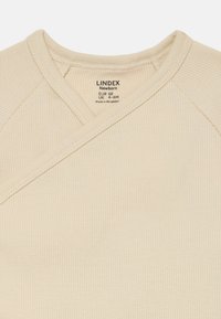 Beige ribbet nyfødt omslagstopp med krysshalsdesign. Lappen viser merke og størrelse. Myk tekstur.