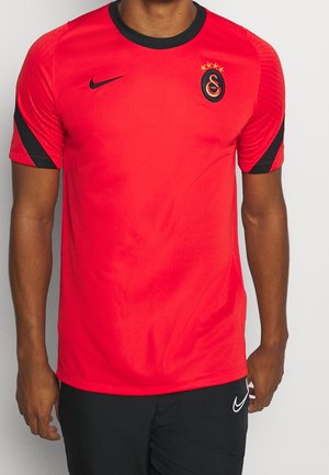 Hombre con una camiseta deportiva roja y negra con un logo de equipo y pantalones cortos negros de Nike, de pie sobre un fondo blanco.