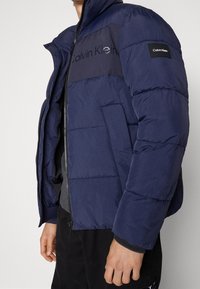 Marinblå puffjacka med kviltad textur, dragkedja och hög krage. Har en kontrasterande svart panel och logotyp på bröstet och ärmen.