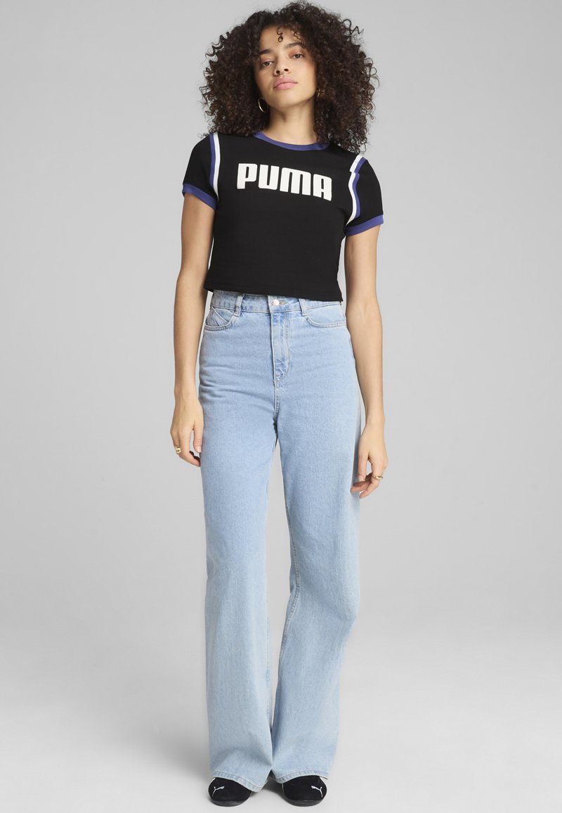 Camiseta negra corta con acentos en azul y blanco y logo de "PUMA". Jeans de tiro alto en azul claro con pantalones acampanados. Modelo lleva calcetas negras.