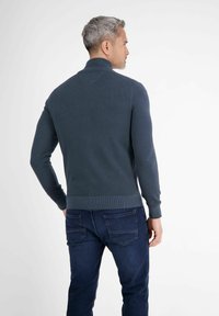 Mężczyzna z siwymi włosami, ubrany w ciemnoniebieski sweter z dzianiny i ciemne dżinsy, stoi tyłem na jednolitym białym tle.