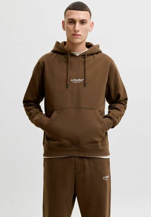 Jeune homme portant un sweat à capuche marron JJ Studios et un pantalon de jogging assorti, les mains dans la poche du sweat, sur un fond uni.