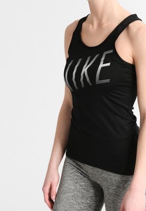 Kobieta ubrana w czarny bezrękawnik z białym napisem "NIKE" i szare legginsy sportowe, stojąca na jednolitym jasnym tle.