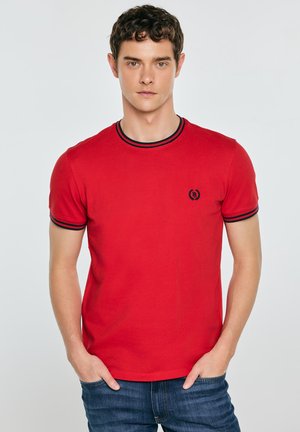 Noor mees lühikeste lokkis juustega, kes kannab punast lühikeste varrukatega särki musta äärega ja väikese logoga, seisab käed sinistes teksapükste taskutes.