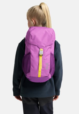 Laps, kellel on blond hobusesaba, kannab musta jope ja lilla seljakotti kollase rihmaga, vaadates selja tagant tühja tausta ees.