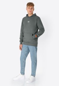 Jonge man in een grijze hoodie, lichte spijkerbroek en witte sneakers, staand met één hand in de zak van de hoodie.