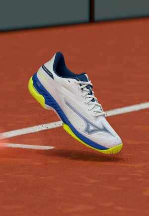 Zapatilla de tenis blanca con suela azul y amarilla, texturizada con un patrón para agarre. Cordones y cuello acolchado, suspendida sobre una cancha de arcilla.