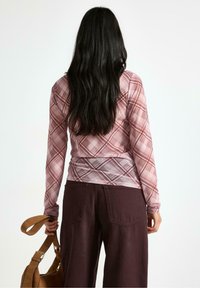 Langærmet top i lyserød ternet mønster, med et gather-design i taljen. Stylet med brede bukser og en brun skuldertaske.