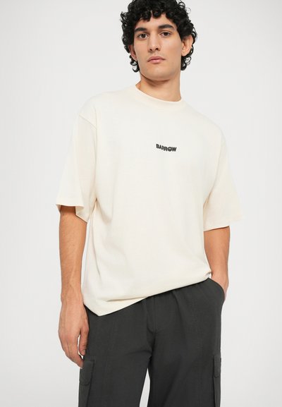 T-shirt oversize crema in cotone con maniche corte, con un piccolo logo nero centrato sul petto. Indossata con pantaloni cargo scuri.