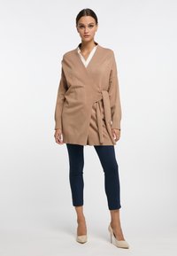 Beige wikkelcardigan van een zachte gebreide stof, voorzien van lange mouwen, een strikdetail in de taille en geribbelde manchetten; gecombineerd met marineblauwe leggings.