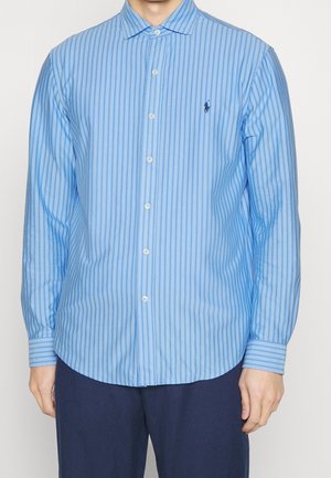 Homme portant une chemise bleu clair à boutons avec des rayures verticales et un pantalon bleu marine foncé, debout devant un fond uni.