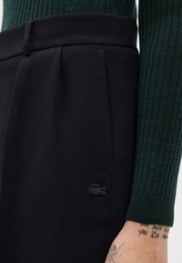 Gros plan sur une personne portant un pantalon noir texturé avec un petit logo crocodile brodé et un pull à manches longues en maille côtelée de couleur vert foncé.