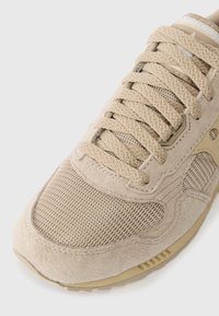 Sapatilha atlética bege com uma combinação de materiais em camurça e rede, atacadores redondos, superfície texturizada e sola acolchoada.