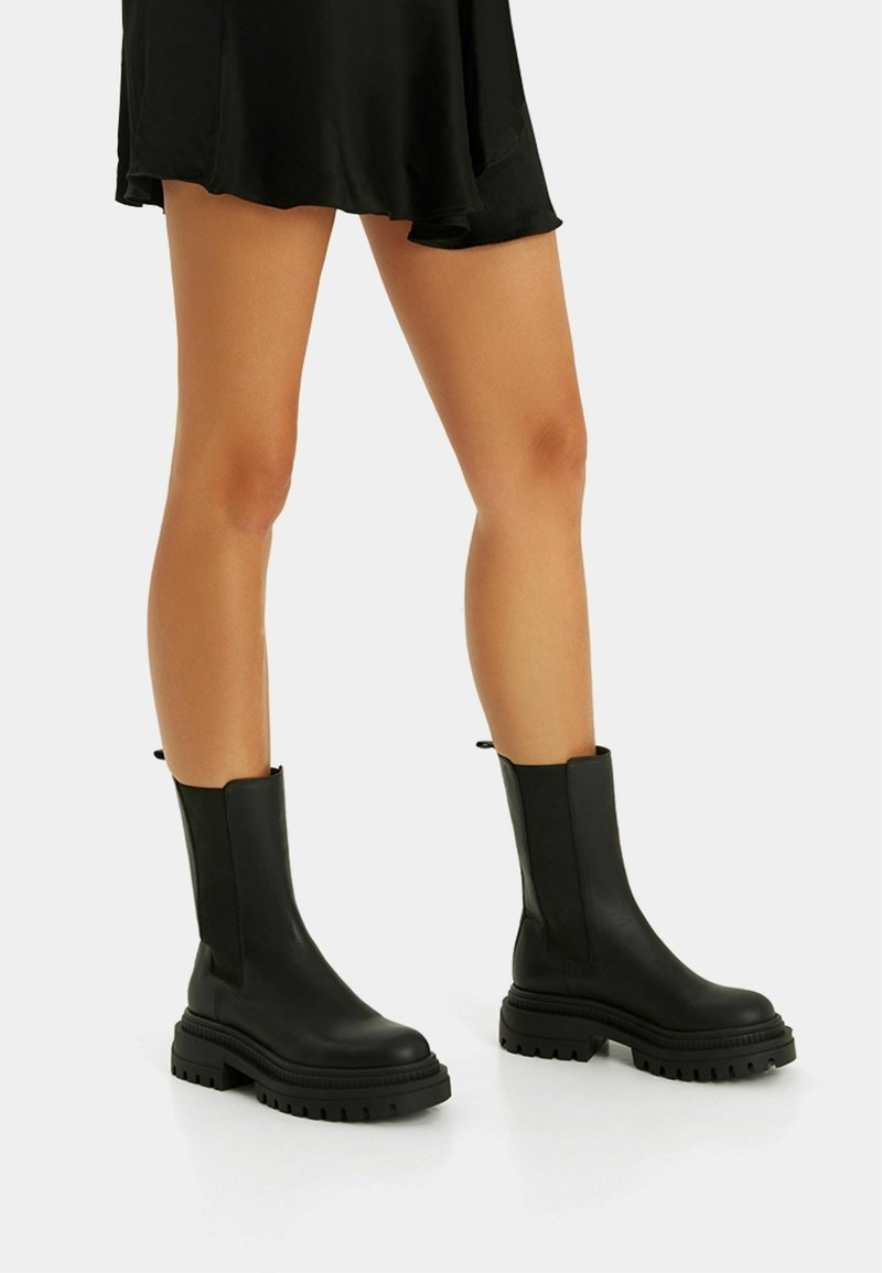 Botas negras de tobillo hechas de cuero suave con una suela gruesa. Cuentan con paneles elásticos en los laterales y tiradores para facilitar el uso.