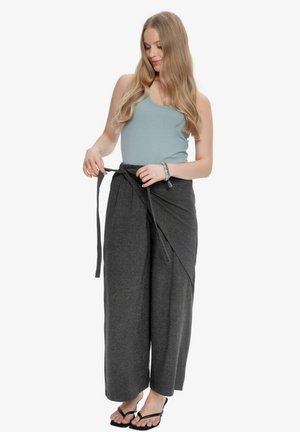 Kobieta z długimi blond włosami, ubrana w jasnoniebieski top na ramiączkach i ciemnoszare szerokie spodnie, wiążąca pasek w talii, nosząca sandały.