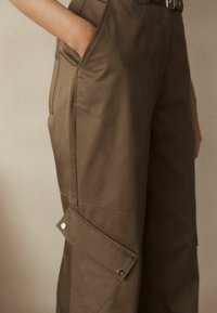 Pantaloni cargo maro cu o textură netedă, buzunare laterale mari și detalii metalice argintii. Ajustați la talie, cu un croi lejer pe picioare.