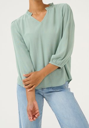 Žena v světlém zeleném dlouhém rukávu s volánovým límcem a světle modrých džínách, držící jednu paži druhou rukou před sebou.